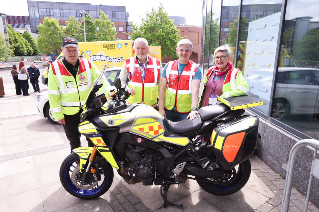 Four adults in hi-vis jackets stand next to a yellow emergency motorcycle parked on a city pavement. A reflective glass building is behind them, with another emergency vehicle partially visible to the right.