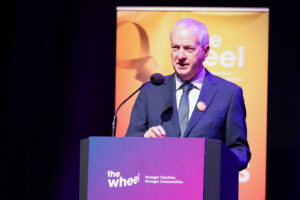 A man in a suit speaks at a podium with the Wheel logo and slogan. A yellow and orange banner is in the background. The setting appears to be a formal event or conference.