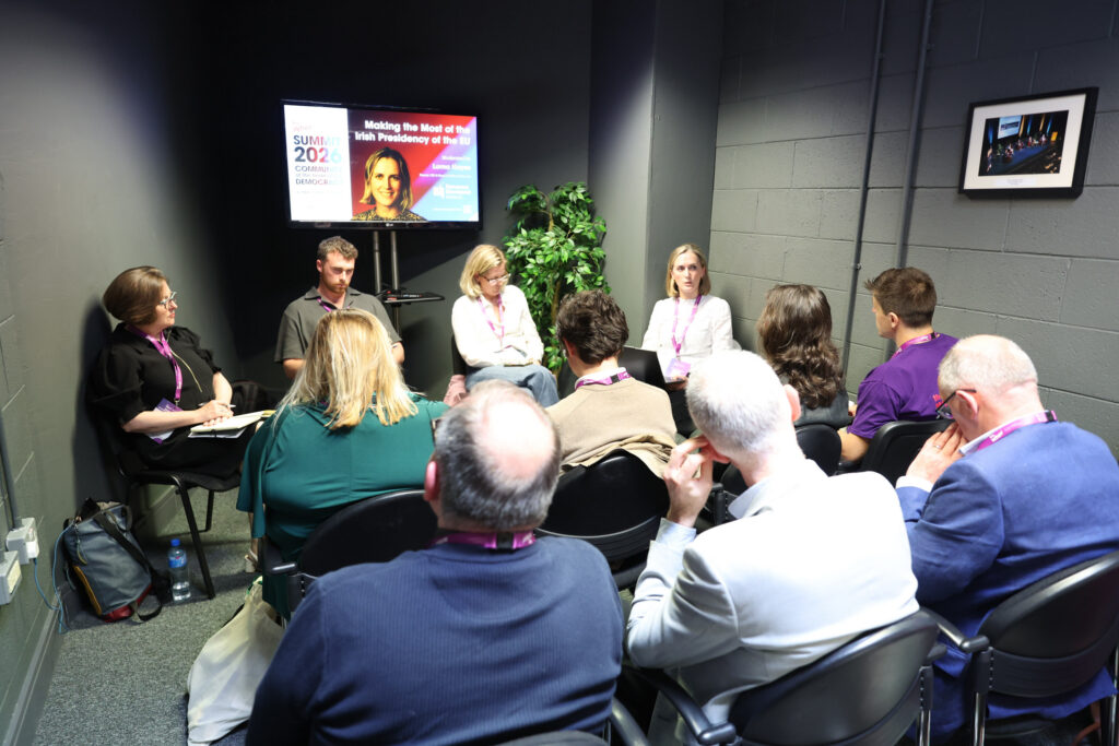 A small group of people sit in chairs facing a panel of five speakers in a room with gray walls. A presentation slide on a screen displays the title Making the Most of the Irish Presidency of the EU..
