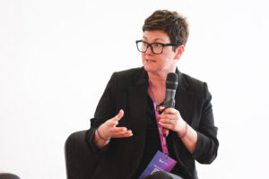 A person with short brown hair and glasses, dressed in a black blazer, sits and speaks into a handheld microphone, gesturing with one hand while holding a folder on their lap. A purple lanyard hangs around their neck.