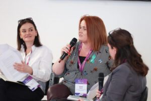 Three women sit and talk during a panel discussion. Two hold papers, all wear conference badges and have microphones. They are engaged in conversation, one woman speaking while the others listen attentively.