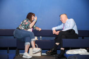 A woman and a man sit on blue benches against a blue wall, engaged in conversation. The woman gestures with her hand, and both have bags on the bench beside them.