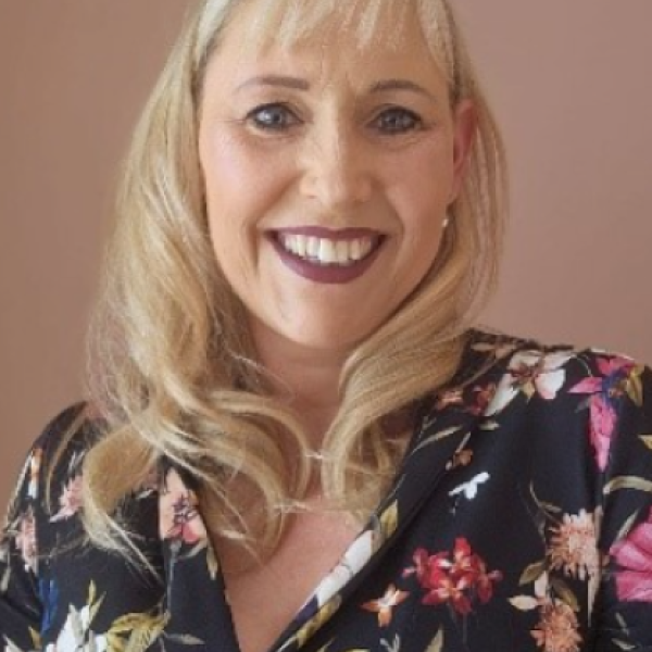A woman with blonde hair and a deep side part is smiling. She wears a black floral blouse and dark lipstick. The background is plain and light brown.
