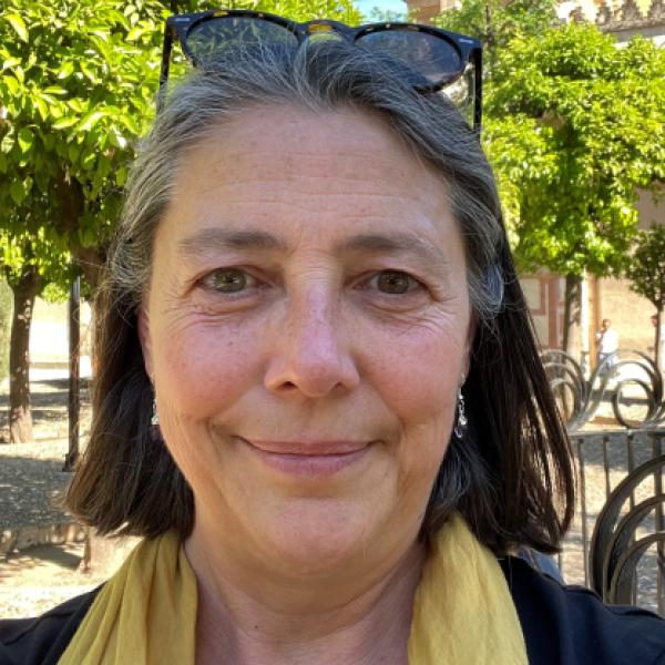 A woman with gray and dark hair is outdoors, smiling at the camera. She is wearing a yellow scarf and glasses on her head. Trees and a building are visible in the background.