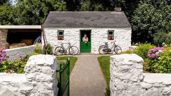 A person stands at the green door of a small white stone cottage with two bicycles outside. The garden has colorful flowers, and a stone path leads from the gate to the entrance. Trees are in the background.