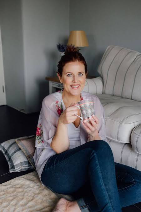 A woman with red hair sits on a cushion in a living room, holding a mug and smiling at the camera. She wears a light purple blouse and jeans. A striped couch and a lamp are in the background.