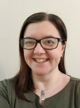 A person with long brown hair and glasses smiles at the camera. They are wearing a green top and a pendant necklace. The background is a plain, light-colored wall.