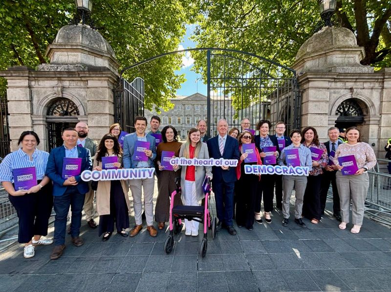 A large group of people stands in front of an ornate iron gate, holding purple pamphlets that read Community at the heart of our democracy as part of a Budget 2027 campaign by The Wheel.