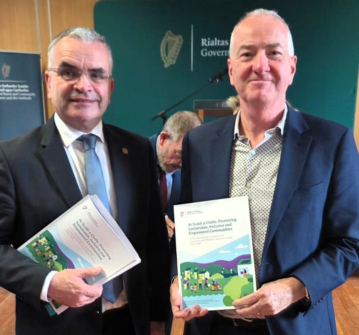 Two men in suits standing side-by-side, each holding a copy of a report titled Ar Scáth a Chéile: Promoting Sustainable, Inclusive and Empowered Communities.