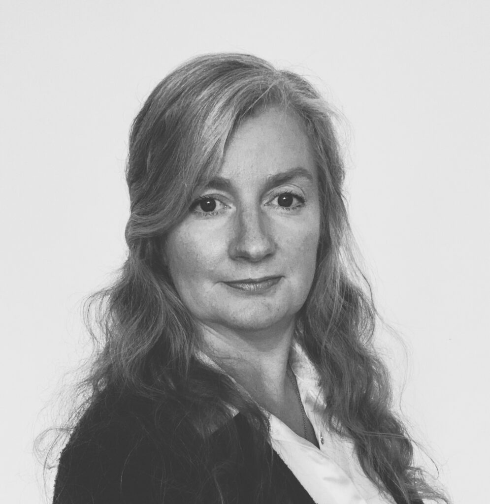 A professional headshot of a woman with long, fair hair wearing a dark blazer over a white shirt.