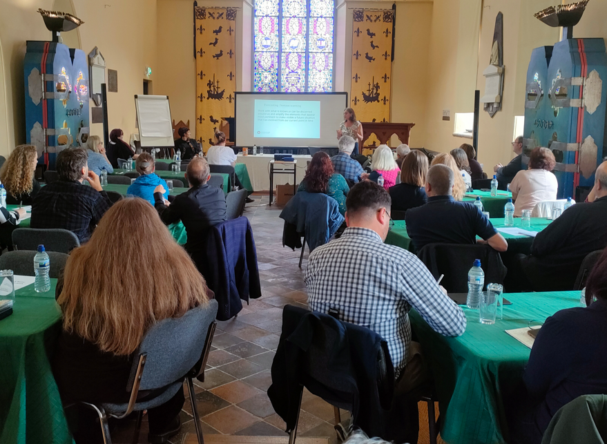 A woman stands at the front of a room giving a presentation to a group of seated attendees at tables covered in green cloths.